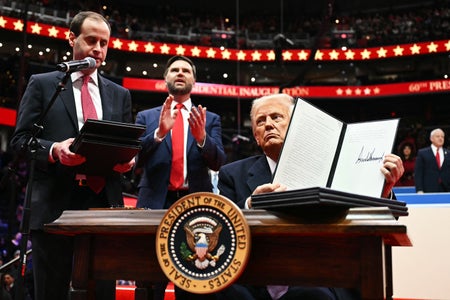 US President Donald Trump sits at a desk with the presidential seal displayed on the front whild holding an executive order announcing the US withdrawal from the Paris Agreement, he just signed during the inaugural parade inside Capital One Arena, in Washington, DC, on January 20, 2025. An aide stands to the left holding a stack of exectuive orders and Vice President J.D. Vance stands behind the desk clapping